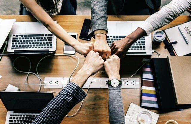 Teamwork and collaboration in action A top-down view of a group of five diverse individuals fist-bumping over a wooden table, surrounded by laptops, notebooks, and other office supplies, symbolizing teamwork and collaboration.