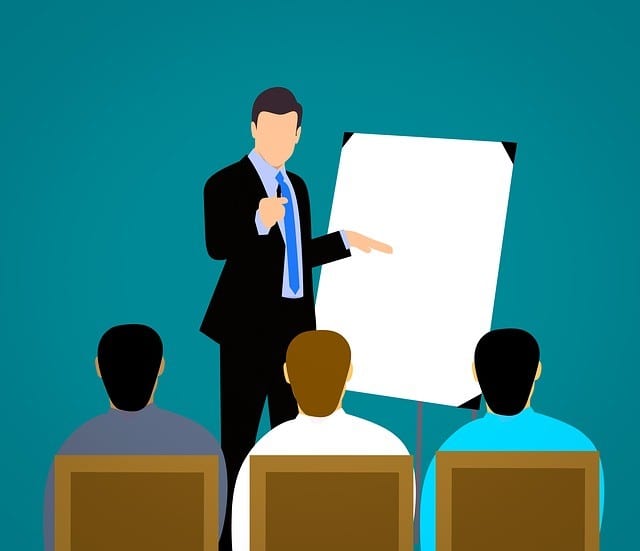 Invest in your people to train them A vector illustration of a business professional in a suit presenting in front of a group of three seated individuals. The presenter is pointing at a blank flipchart while the audience listens attentively