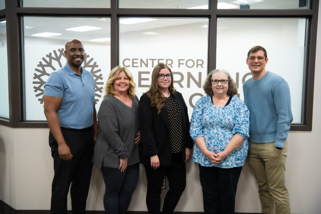 Group photo of five staff members of the Center of Learning and Teaching at the Oklahoma City Community College, standing in front of a large window with 'Center for Learning' text and a snowflake logo visible on the glass. From left to right: a Black man in a light blue polo shirt, a blonde woman in a gray cardigan, a woman with long brown hair wearing glasses and a black polka dot top, a woman with curly hair and glasses wearing a blue floral blouse, and a man in a light blue sweater. They are standing in what appears to be an office or educational facility with white walls and modern lighting.